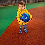 child, yellow_jacket, blue_soccer_ball, playground, outdoor, standing, serious_expression, colorful_ground, fence, jeans, sneakers, young_boy, toy, sport, casual_clothing, daylight, urban, playing, holding, footwear