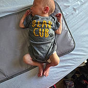 Elliot is registered to the contest to win money with this photo: newborn, baby, sleeping, onesie, pacifier, bed, tissue_box, feet, hands, clothing, infant, cute, resting, indoor, soft, grey, yellow_text, crib_mat, peaceful, child