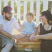 Eveline is registered to the contest to win money with this photo: adult, baby, casual_clothing, crate, dress, family, fence, glasses, grass, happy, man, nature, outdoor, shorts, sitting, smiling, sunlight, tattoos, toy_car, woman