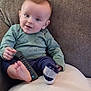 baby, child, remote_control, couch, green_shirt, dinosaur_pattern, navy_pants, sock, barefoot, pillow, indoor, smiling, face, person, seated, cozy, soft_lighting, curious, young_child, casual
