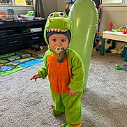 Koleson is registered to the contest to win money with this photo: child, toddler, dinosaur_costume, costume, pacifier, indoor, living_room, carpet, toy, playroom, inflatable, green, orange, standing, portrait, cute, window, playmat, basket, furniture
