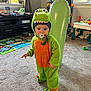 child, toddler, dinosaur_costume, costume, pacifier, indoor, living_room, carpet, toy, playroom, inflatable, green, orange, standing, portrait, cute, window, playmat, basket, furniture