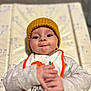Jackson is registered to the contest to win money with this photo: baby, hat, bib, hands, face, infant, child, lying_down, patterned_mat, knit_hat, orange_trim, curious_expression, clothing, cute, person, indoor, closeup, soft_lighting, skin, eyes