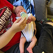 Michael is registered to the contest to win money with this photo: baby, bottle, feeding, onesie, couch, blanket, adult, hand, red_shirt, jeans, footwear, crocs, cushion, indoor, relaxing, infant, person, comfort, casual, peaceful