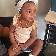Mia Carolina is registered to the contest to win money with this photo: toddler, child, smiling, headband, white_bow, sitting, indoors, shorts, socks, necklace, curly_hair, cute, happy, person, furniture, table, black_surface, skin_tone, casual, home