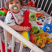 Charly participe au concours pour gagner de l'argent avec cette photo : toddler, crib, pacifier, toy, stacking_cone, plush_toy, socks, bib, curly_hair, indoor, floor_tiles, child, playtime, colorful, red, green, blue, yellow, white, polka_dot