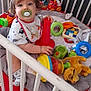 toddler, crib, pacifier, toy, stacking_cone, plush_toy, socks, bib, curly_hair, indoor, floor_tiles, child, playtime, colorful, red, green, blue, yellow, white, polka_dot