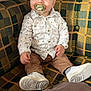 toddler, child, pacifier, white_shirt, brown_pants, white_sneakers, couch, plaid_pattern, indoor, pillow, happy, curious, sitting, person, footwear, home, casual_clothing, portrait, young_child, smiling