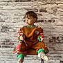 child, toddler, sitting, stool, brick_wall, indoor, portrait, colorful_clothing, socks, paintbrush, looking_away, brown_outfit, patterned_sleeves, short_hair, curious_expression, studio_style, cozy, young_child, seat, casual