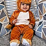 baby, child, costume, lion_costume, smiling, cute, indoors, couch, pillows, patterned_pillows, plush, happy, seated, soft_texture, small_hands, footwear, headgear, portrait, cozy, warm_colors