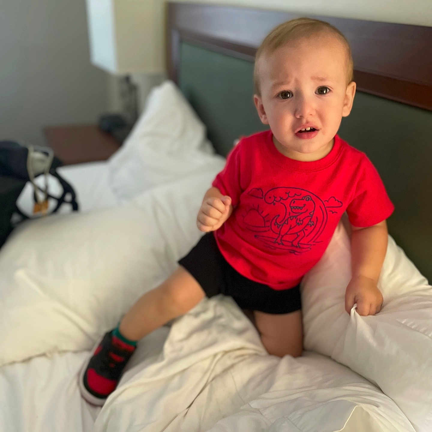 Othrin joined the competition — help win amazing prizes! bed, black_shorts, blanket, casual_clothing, child, confused_expression, dinosaur_print, footwear, furniture, headboard, indoor, lamp, person, pillow, red_tshirt, room, shoe, toddler, white_bedding, young_child