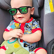 Adam is registered to the contest to win money with this photo: baby, child, sunglasses, car_seat, plush_toy, dinosaur_toy, smiling, infant, cute, clothing, seat_belt, red_trim, happy, indoors, person, toy, hand, legs, head, face