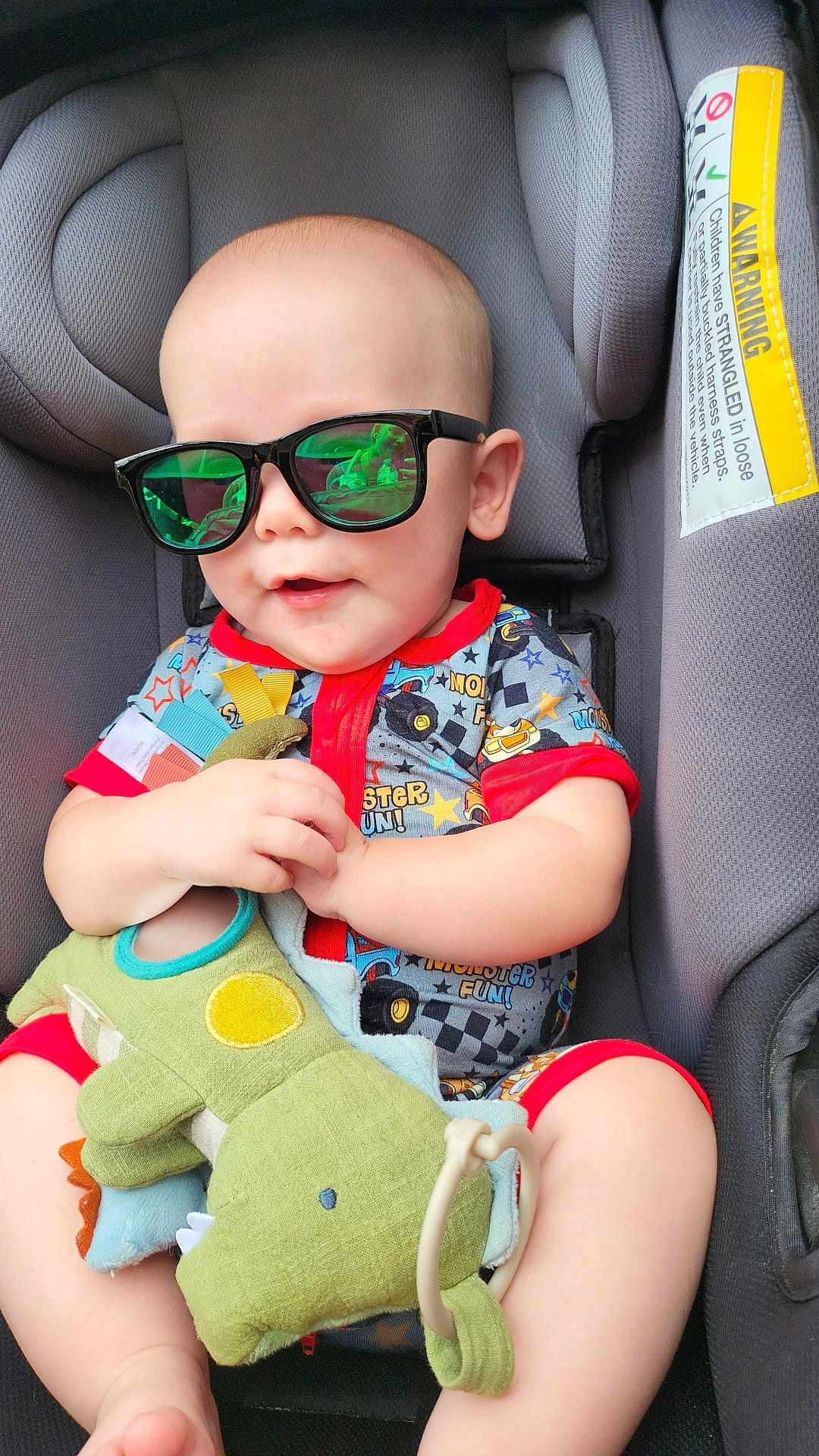 Adam is registered to the contest to win money with this photo: baby, child, sunglasses, car_seat, plush_toy, dinosaur_toy, smiling, infant, cute, clothing, seat_belt, red_trim, happy, indoors, person, toy, hand, legs, head, face
