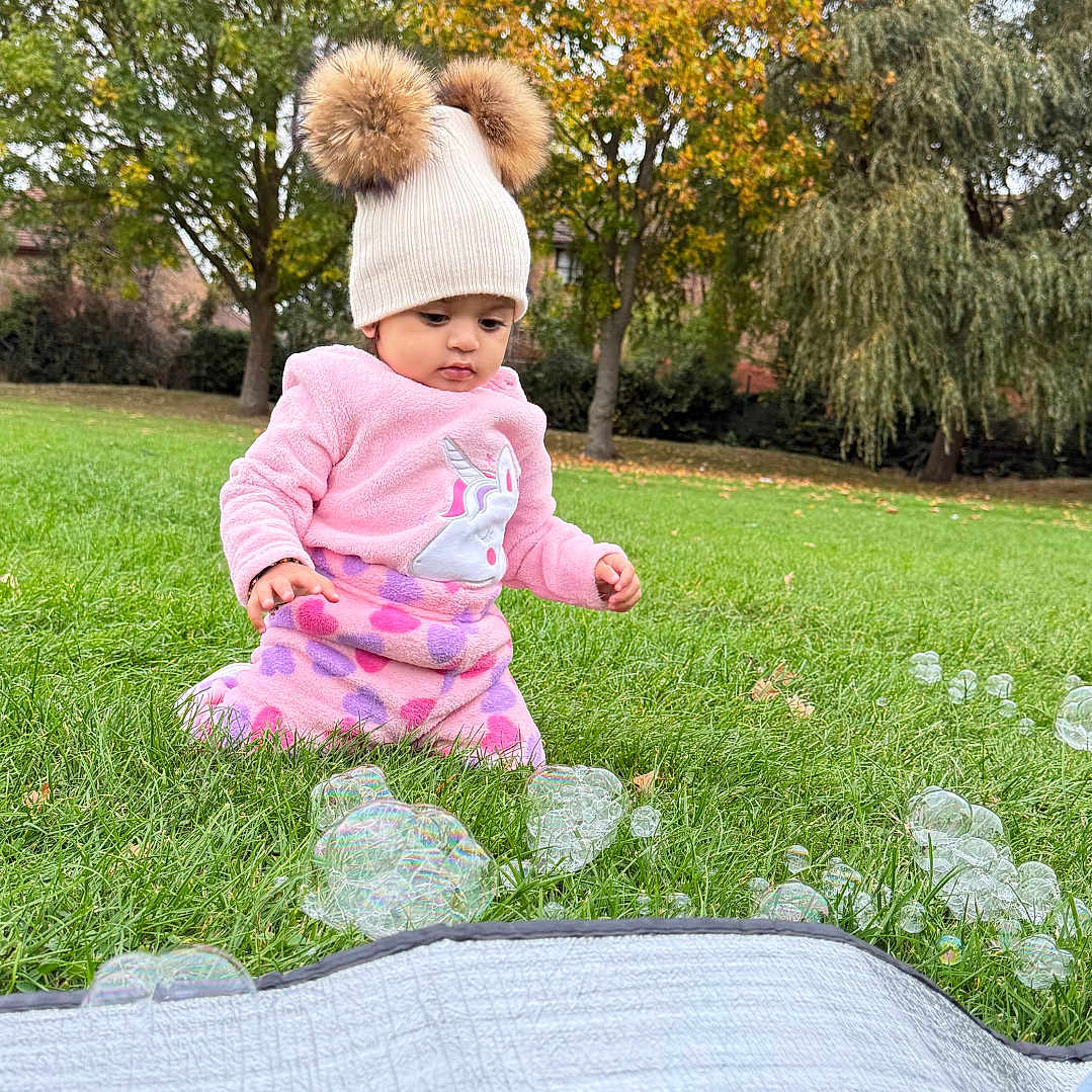 Diara joined the competition — help win amazing prizes! autumn, bubbles, child, curious, cute, daylight, fall, grass, greenery, hat, nature, outdoor, park, pink_clothing, playful, pom_poms, sitting, toddler, trees, young_child