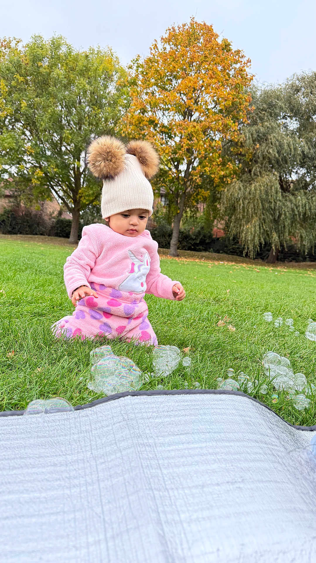 Diara joined the competition — help win amazing prizes! toddler, child, pink_clothing, hat, pom_poms, bubbles, grass, outdoor, autumn, trees, greenery, cute, curious, playful, nature, fall, park, young_child, sitting, daylight