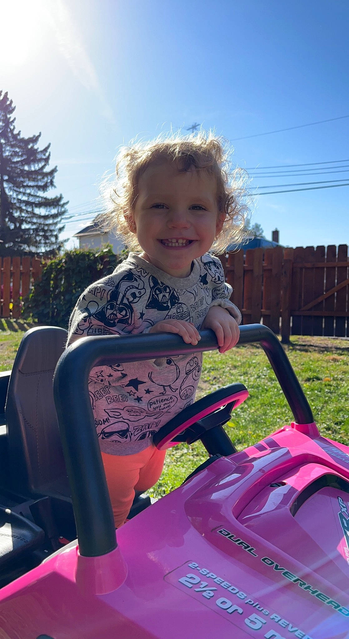 Kinsley is registered to the contest to win money with this photo: automotive_exterior, automotive_tire, bumper, cloud, fun, grass, happy, joy, leisure, magenta, motor_vehicle, person, pink, plant, recreation, riding_toy, sky, smile, t_shirt, toddler