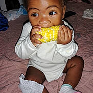 Cyncere is registered to the contest to win money with this photo: baby, child, corn_on_the_cob, eating, bedspread, quilt, socks, white_clothing, hands, face, curly_hair, indoor, cute, infant, munching, mischievous, young_child, portrait, sitting, food