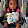 child, toddler, car_seat, lego, toy, shoes, socks, pink_shirt, seatbelt, safety, indoor, curly_hair, cute, young_child, person, footwear, holding, box, expression, portrait