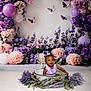 baby, child, flower_arrangement, purple_flowers, pink_flowers, butterflies, basin, indoor, smiling, portrait, decor, greenery, cute, toddler, sitting, white_background, nature_theme, playful, floral, headwear