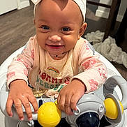 Delilah is registered to the contest to win money with this photo: baby, child, smiling, headband, bow, bib, toy, walker, hands, face, indoor, floor, blanket, clothing, person, happy, cute, infant, home, playtime
