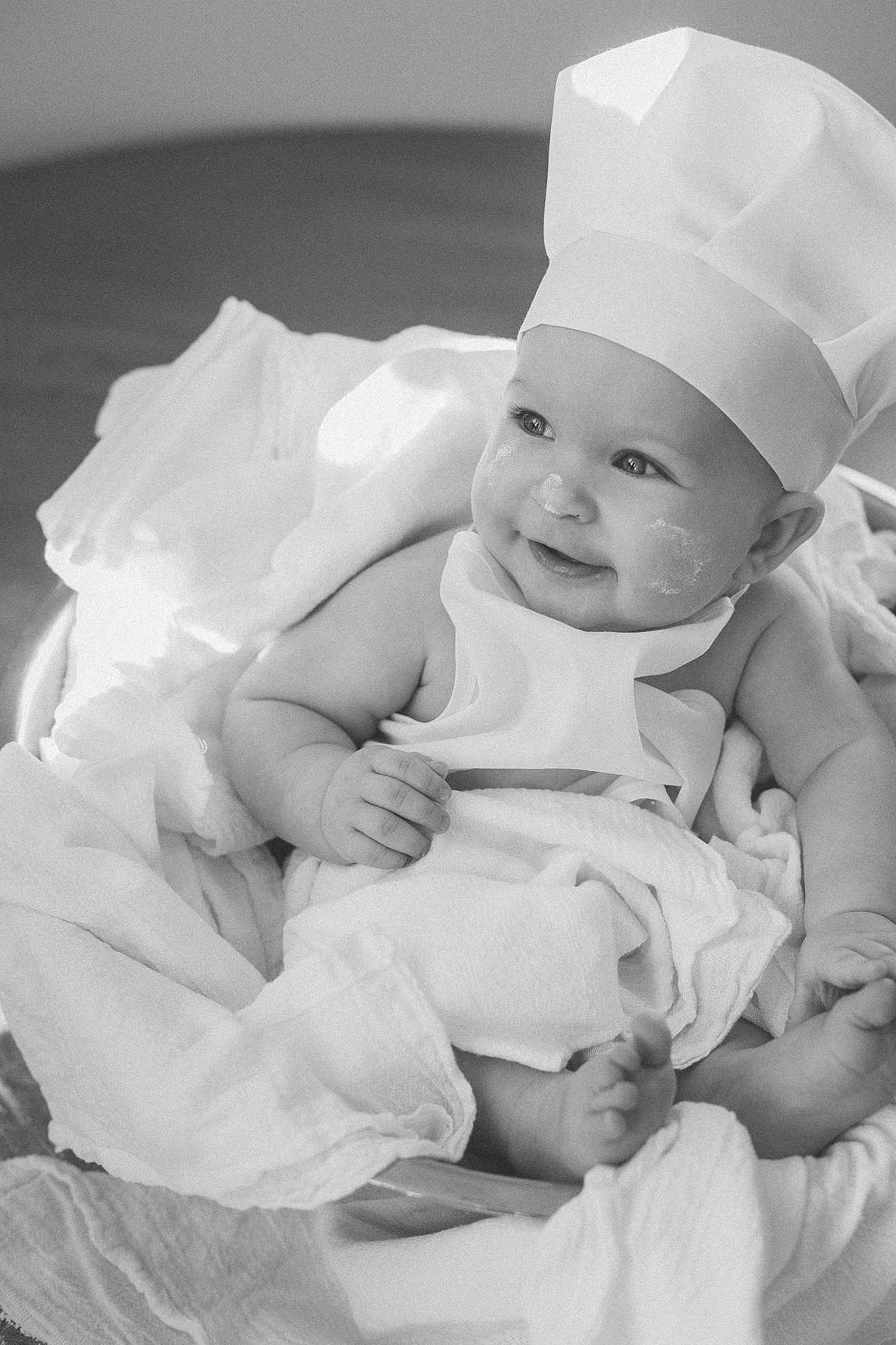 Charlotte joined the competition — help win amazing prizes! baby, infant, child, chef_hat, costume, smile, face, hands, feet, blanket, cloth, bowl, cooking, flour, cute, portrait, indoors, black_and_white, happy, playful