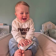 Mahon a rejoint le concours — aidez-le/la à gagner de superbes lots ! baby, infant, smiling, laughing, sitting, nursery, remote_control, bare_feet, jeans, hoodie, rosy_cheeks, portrait, child, indoor, wall, crib, soft_toy, decorative_sign, happy, hands