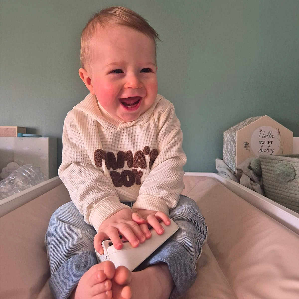 Mahon a rejoint le concours — aidez-le/la à gagner de superbes lots ! baby, bare_feet, child, crib, decorative_sign, hands, happy, hoodie, indoor, infant, jeans, laughing, nursery, portrait, remote_control, rosy_cheeks, sitting, smiling, soft_toy, wall