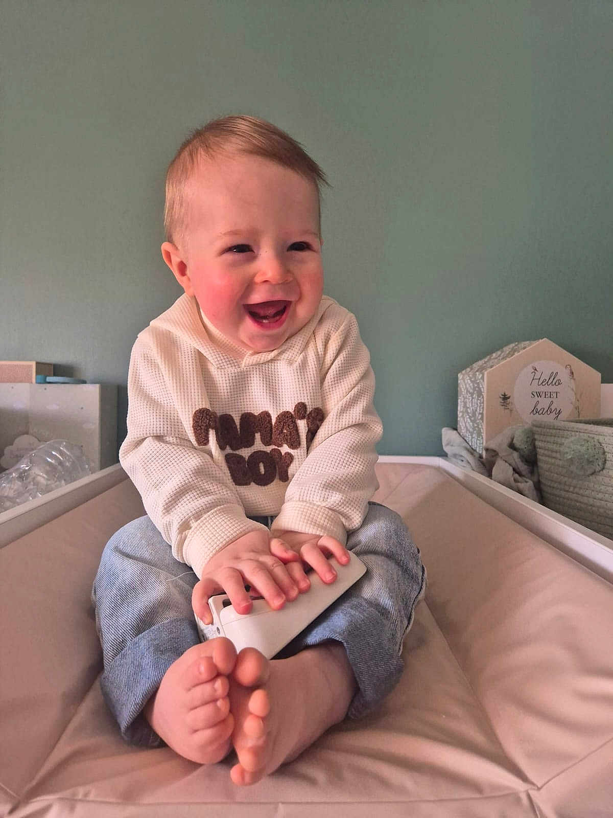 Mahon a rejoint le concours — aidez-le/la à gagner de superbes lots ! baby, infant, smiling, laughing, sitting, nursery, remote_control, bare_feet, jeans, hoodie, rosy_cheeks, portrait, child, indoor, wall, crib, soft_toy, decorative_sign, happy, hands