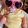 baby, child, pacifier, sunglasses, heart_shaped, yellow_onesie, nike_logo, indoor, cute, infant, hand, face, clothing, person, fashion, glasses, closeup, portrait, seated, adorable