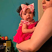 Journee is registered to the contest to win money with this photo: baby, woman, holding, pink_dress, headband, bow, arm, indoor, person, curly_hair, facial_expression, sleeveless, wall, shelf, bottle, toy, colorful, casual, closeup, portrait