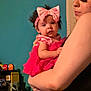 baby, woman, holding, pink_dress, headband, bow, arm, indoor, person, curly_hair, facial_expression, sleeveless, wall, shelf, bottle, toy, colorful, casual, closeup, portrait