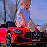 toddler, child, toy_car, red_car, outdoor, grass, flowers, blue_sky, sunny, smiling, shorts, white_shoes, happy, nature, play, daylight, vehicle, small_car, casual_clothing, person