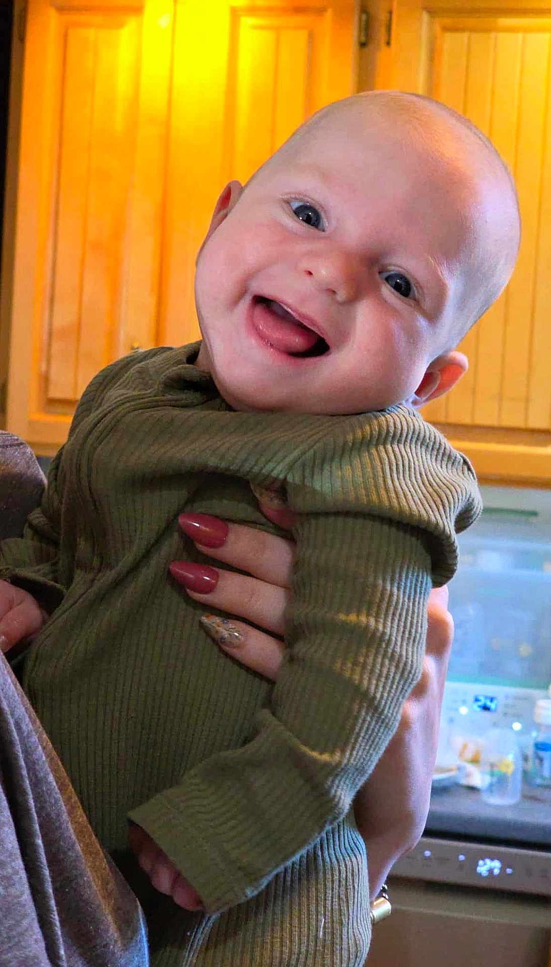Arthur joined the competition — help win amazing prizes! baby, smiling, child, face, person, hand, nail_polish, long_nails, clothing, green_outfit, indoor, kitchen, cabinet, wood, countertop, dishwasher, bottle, happy, portrait, holding