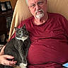 cat, tuxedo_cat, man, person, portrait, indoors, living_room, recliner, seated, lap, pet, companion, gray_fur, white_chest, green_eyes, glasses, beard, red_shirt, family_photos, cozy