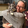 cat, woman, person, portrait, indoors, kitchen, cuddling, holding, gray_cat, pet, glasses, smiling, sweater, whiskers, yellow_eyes, paws, furniture, cabinets, background_person, home