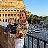 woman, toddler, child, smile, colosseum, ancient_architecture, outdoor, daylight, cityscape, tourist, railing, street, people, car, tree, sky, clothing, braid, holding, travel