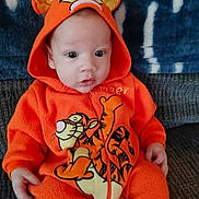 Tommy Isaiah Bernal is registered to the contest to win money with this photo: baby, child, orange_onesie, tigger, hooded_clothing, sitting, couch, blanket, indoor, cute, face, eyes, hands, infant, portrait, warm_clothing, toddler, cozy, soft_fabric, clothing