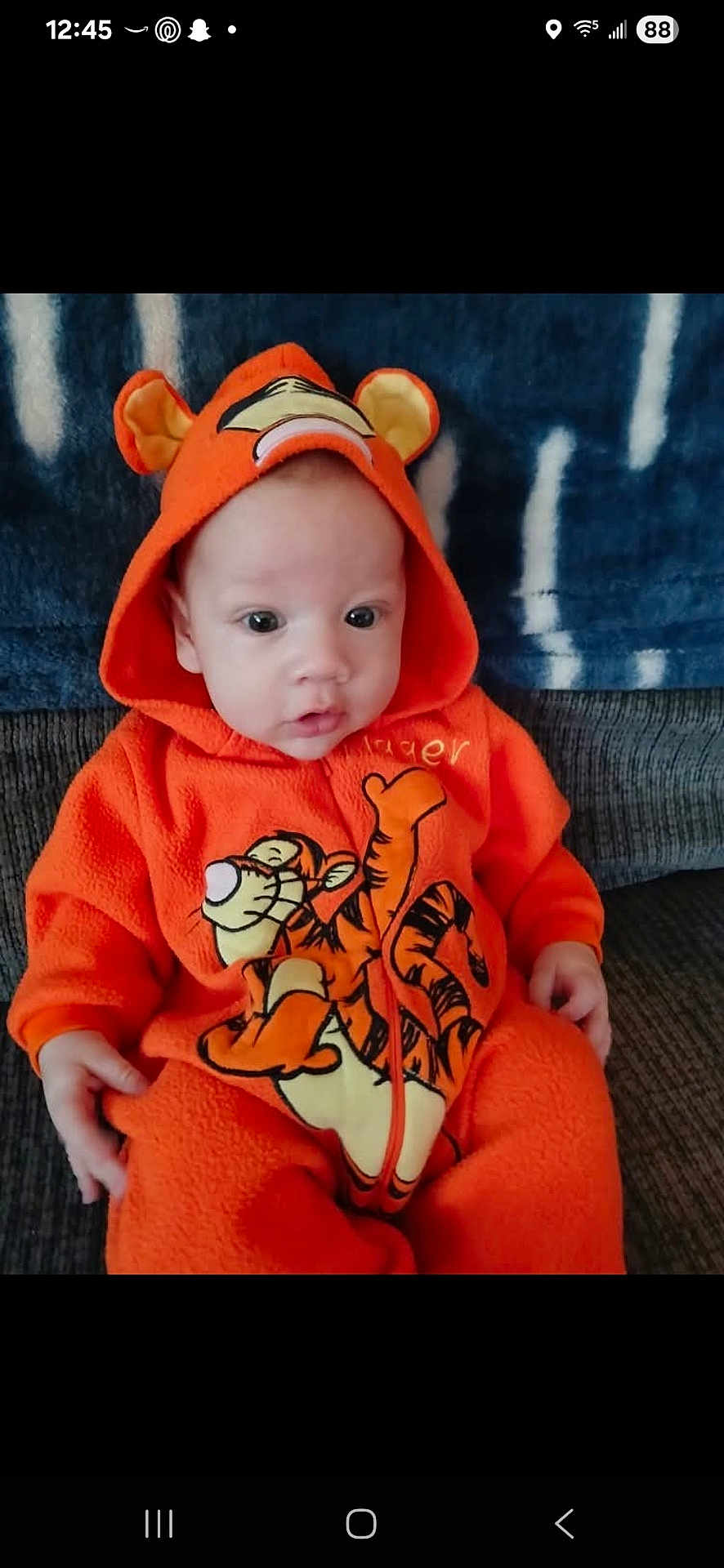 Tommy Isaiah Bernal is registered to the contest to win money with this photo: baby, child, orange_onesie, tigger, hooded_clothing, sitting, couch, blanket, indoor, cute, face, eyes, hands, infant, portrait, warm_clothing, toddler, cozy, soft_fabric, clothing