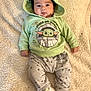 baby, child, hoodie, green_clothing, baby_yoda, star_wars, blanket, white_sneakers, pants, stars_pattern, infant, cute, lying_down, portrait, face, footwear, casual_clothing, soft_texture, indoors, comfortable
