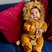 Jude joined the competition — help win amazing prizes! baby, child, lion_costume, plush, couch, red_pillow, blue_couch, toy, cute, smiling, infant, fur_hood, buttoned_outfit, indoor, sitting, portrait, face, hand, footed_pajamas, cozy