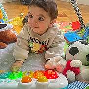 Naël participe au concours pour gagner de l'argent avec cette photo : baby, child, toy, plush_toy, colorful, play_mat, floor, sweatshirt, mickey_mouse, indoor, cute, infant, piano_toy, bright_colors, expression, person, young, crawling, wooden_floor, soft_toys