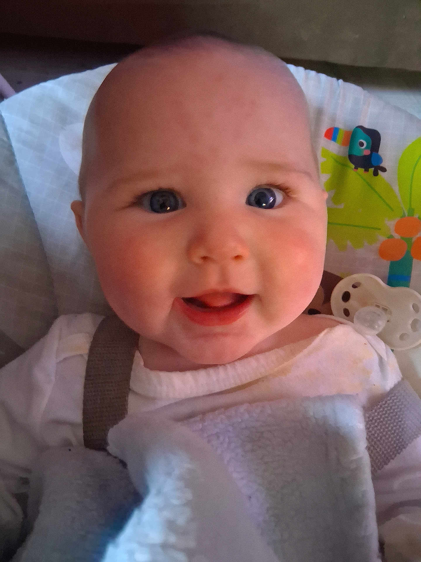 Chloe is registered to the contest to win money with this photo: baby, infant, face, blue_eyes, smile, blanket, white_clothing, pacifier, close_up, cheeks, cute, indoors, child, happy, person, seatbelt, colorful_background, toy, head, portrait