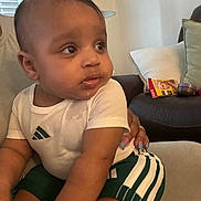 Levi is registered to the contest to win money with this photo: baby, child, infant, person, sitting, clothing, white_tshirt, green_shorts, hand, furniture, couch, pillow, indoor, living_room, skin, face, foot, nail_polish, expression, curious