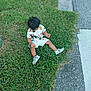 casual_clothing, child, curious, curly_hair, daylight, footwear, grass, greenery, mickey_mouse, nature, outdoor, pavement, person, playful, shirt, sidewalk, sitting, toddler, white_shoes, young_child