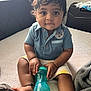 baby, child, sitting, blue_polo, yellow_shorts, sippy_cup, bed, blanket, curly_hair, indoors, cute, chubby, person, infant, hand, foot, window, natural_light, soft_texture, casual