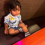 Bhargav Ram is registered to the contest to win money with this photo: toddler, child, sitting, curly_hair, shirt, cartoon, bench, cushion, shoes, indoor, wall, brown, light, table, wood, casual, portrait, young, person, happy