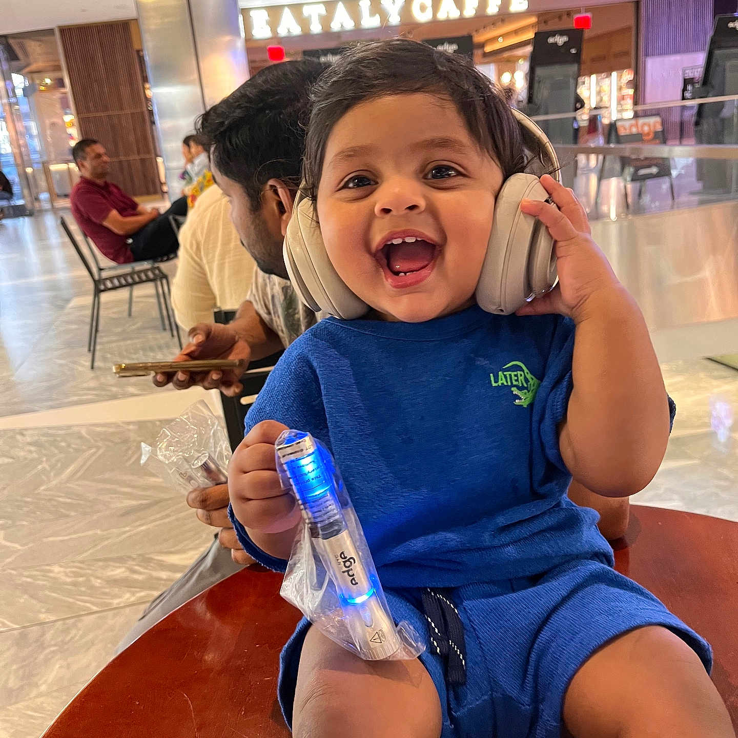 Bhargav Ram is registered to the contest to win money with this photo: background, blue_clothing, cafe, child, face, floor, hair, hand, happy, headphones, indoor, laughing, light, people, person, sitting, smile, table, toddler, toy