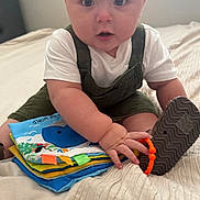 Francisco is registered to the contest to win money with this photo: baby, child, indoor, bed, soft_book, overalls, white_shirt, toy, curious, sitting, hand, footwear, blanket, window, natural_light, cute, infant, playing, person, expression