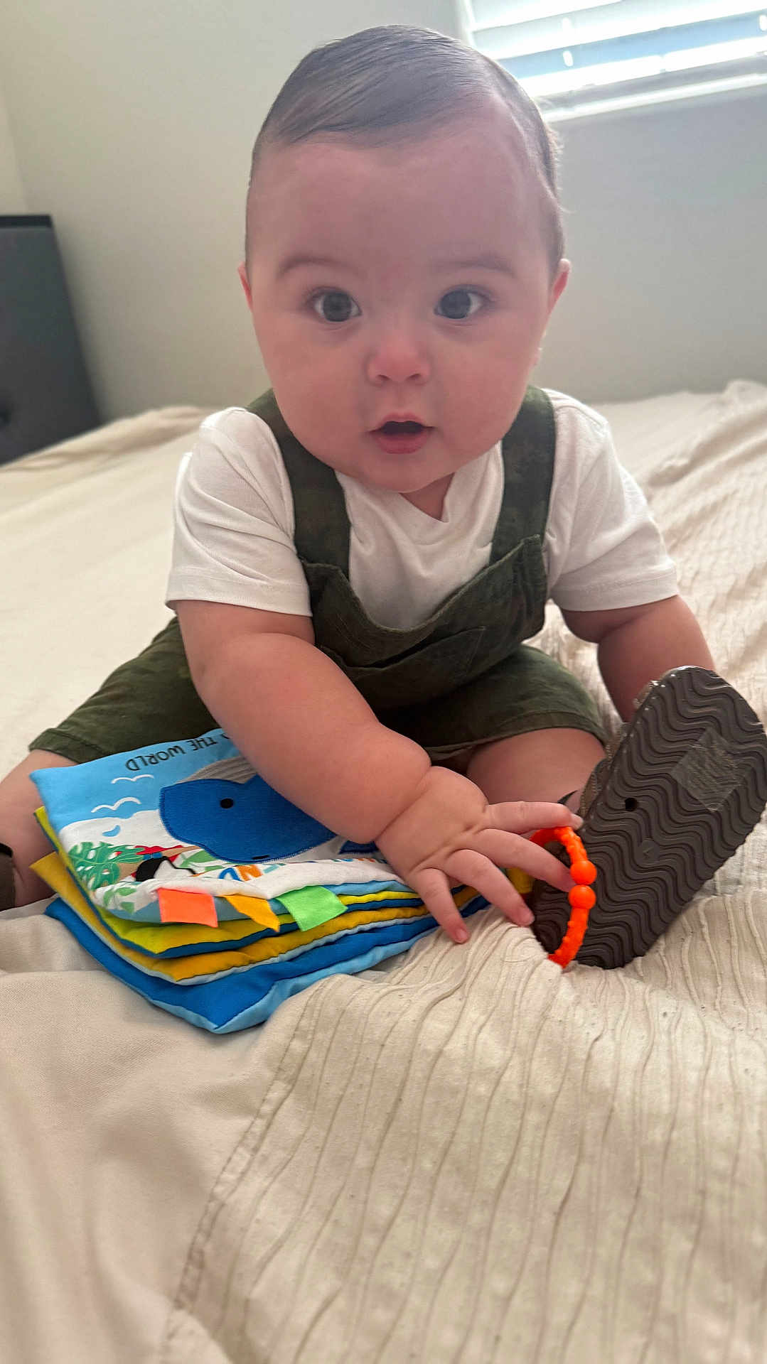 Francisco is registered to the contest to win money with this photo: baby, child, indoor, bed, soft_book, overalls, white_shirt, toy, curious, sitting, hand, footwear, blanket, window, natural_light, cute, infant, playing, person, expression