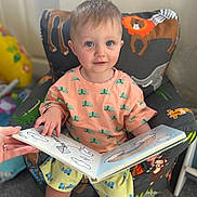 Cobie is registered to the contest to win money with this photo: toddler, child, book, reading, chair, animal_print, indoor, colorful, cute, smiling, person, hand, carpet, clothing, shorts, shirt, toy, illustration, playful, cozy